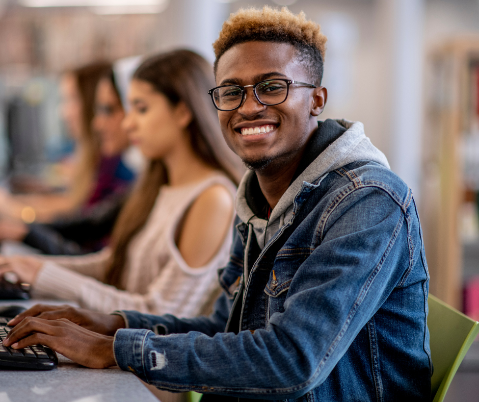 A male student of colour is sitting at desk, smiling. He has bleached blond hair and is wearing a denim jacket