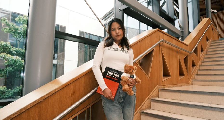 Student standing on the steps of the Streatham Campus Forum building holding a Graduate Employers rankings books and a University of Exeter branded teddy bear